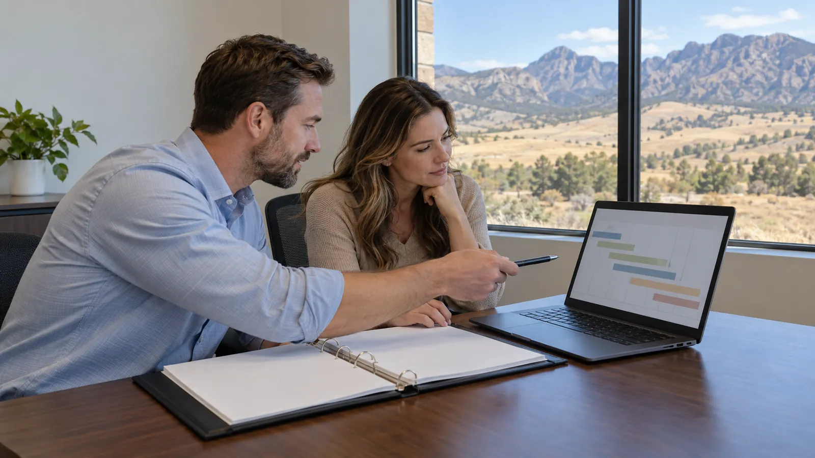 Two people review a contract binder and a schedule chart on a laptop in an office with dry foothills outside
